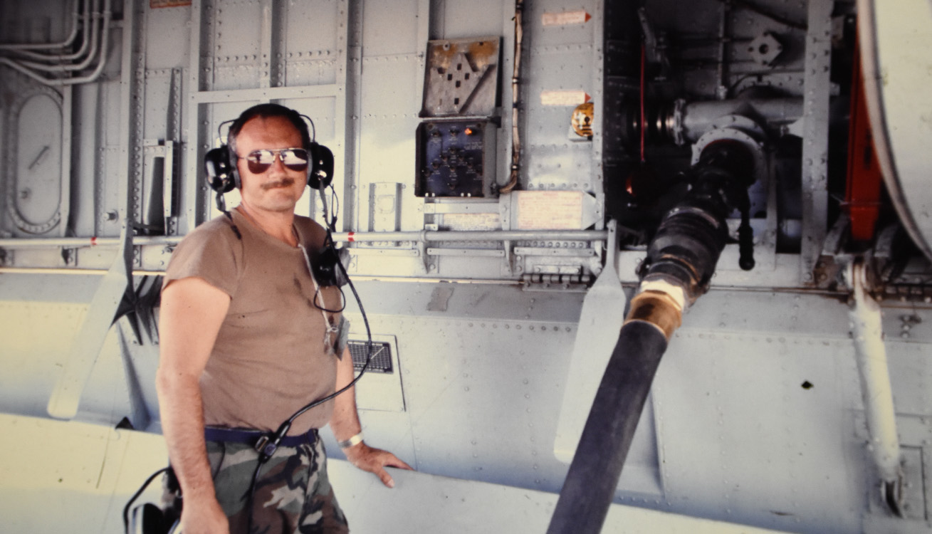 service member smiles at the camera while a fuel hose is hooked up to an aircraft underbelly